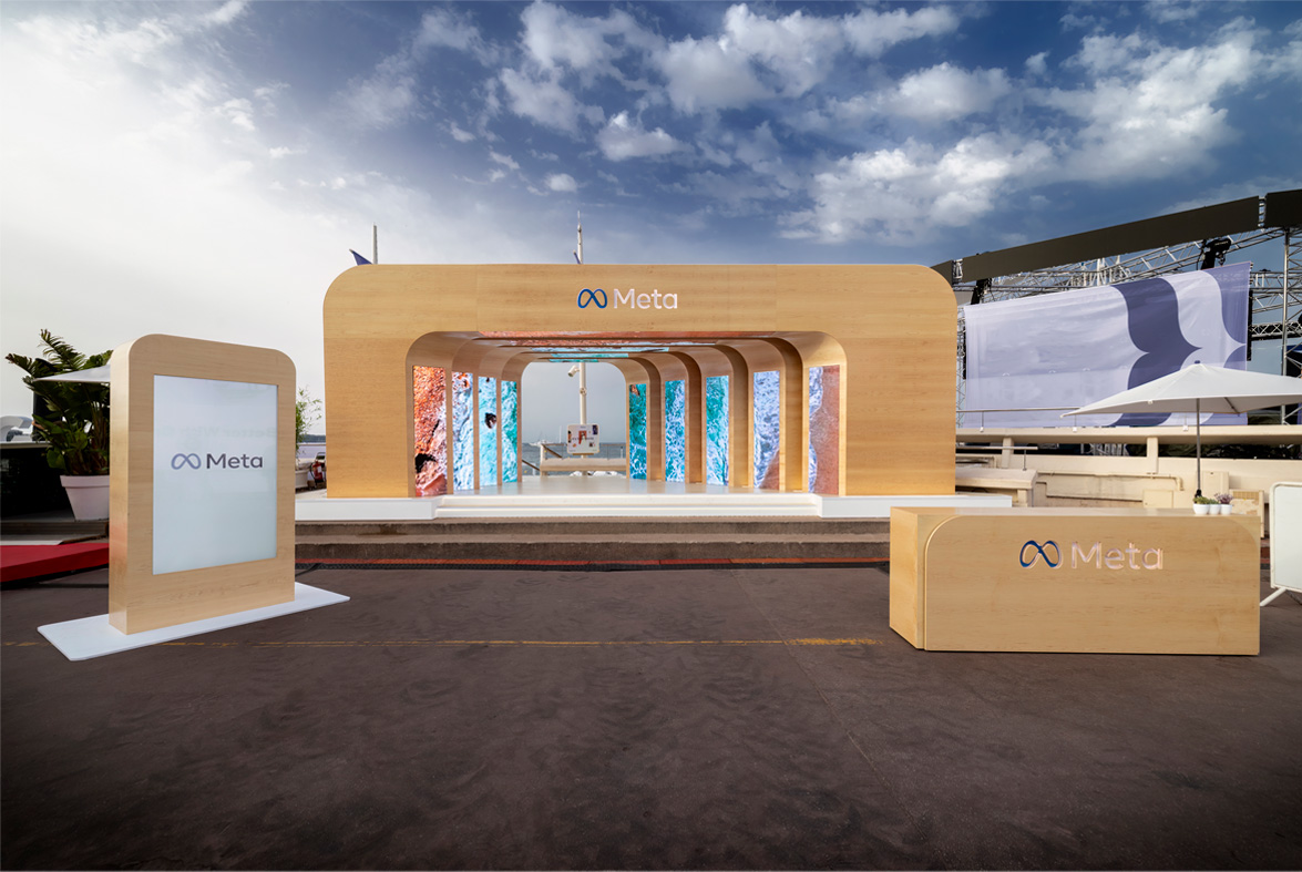 A large, wooden, organic shaped entrance is erected outdoors before a beach. The wooden entrance has the Meta logo engraved into the top center. The entrance forms a tunnel to the beach with screens on both sides of the tunnel revealing different beach textures. Next to the entrance is a large screen that says Meta and a wooden table.